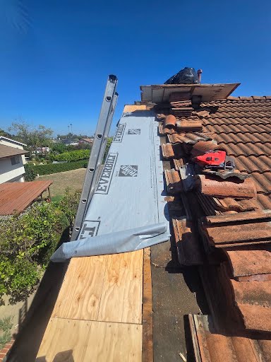Cerritos tile roof during leak repair: rows of clay barrel tiles lifted and stacked on the slope, new plywood sheathing at the eave with gray synthetic underlayment rolled out over it, and a ladder set at the roof edge.