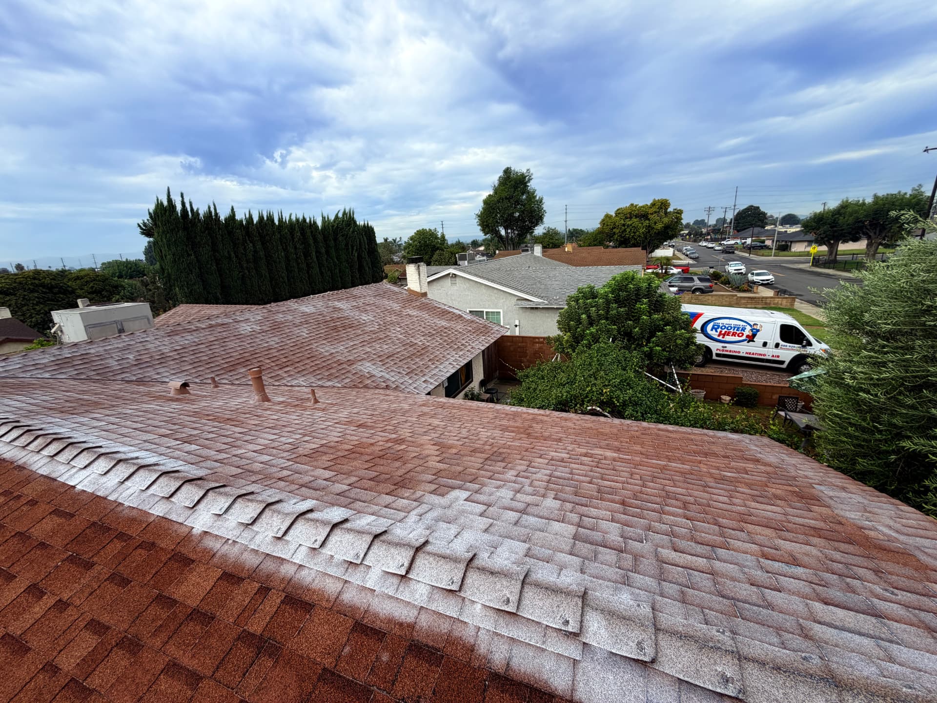Chino roof mid-restoration: reddish-brown asphalt composition shingles with the rejuvenating treatment freshly applied across the front slope and ridge caps, the upper slope still awaiting its coat, and a street of single-story homes in the background.